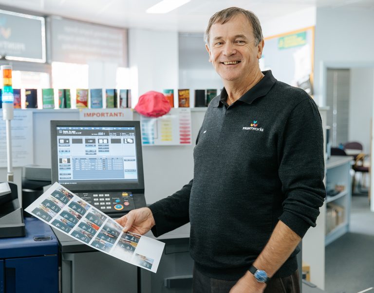 Man holding print samples beside a Konica Minolta printer in an office