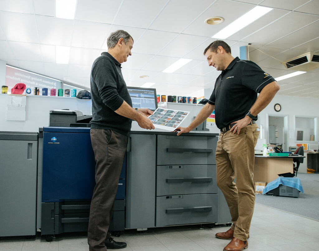 2 men looking down at a printed sheet of paper in front of a production printer